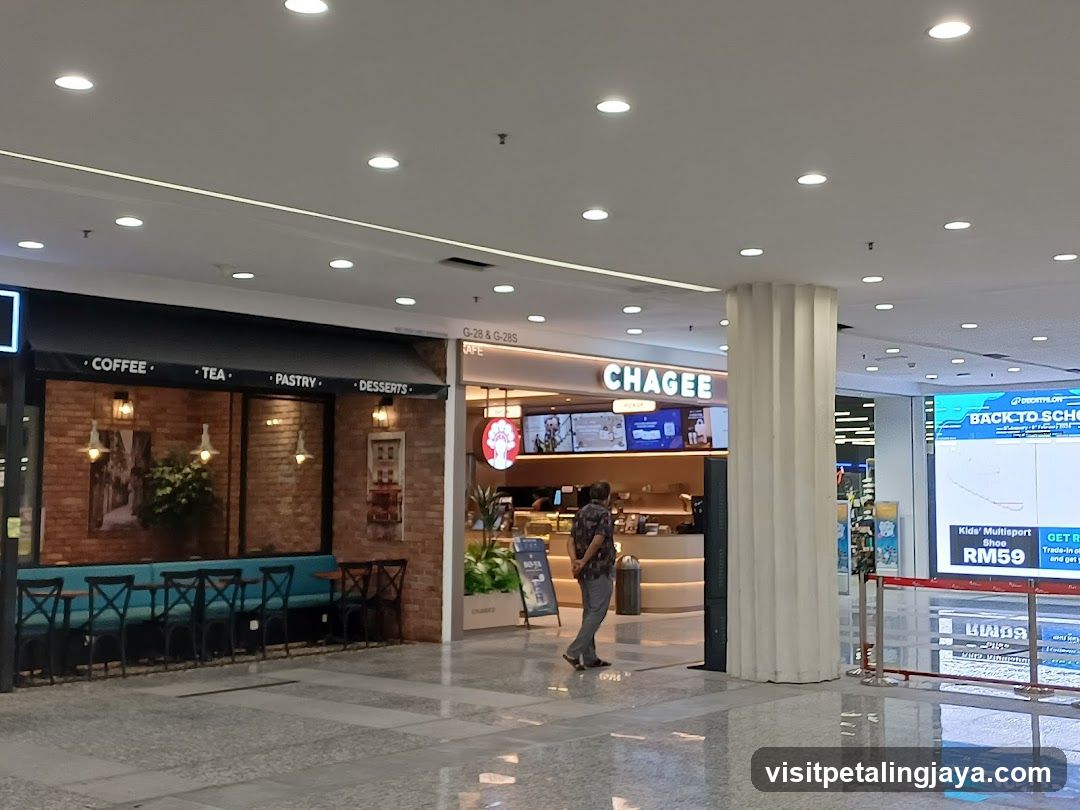A cafe storefront with a black awning displaying the words "COFFEE", "TEA", "PASTRY", and "DESSERTS". The facade features brick walls, large windows, and outdoor seating with blue benches and black chairs. Adjacent to the cafe is a store with the prominent signage "CHAGEE". Further to the right, a large digital screen displays a "DECATHLON BACK TO SCHOOL" advertisement.