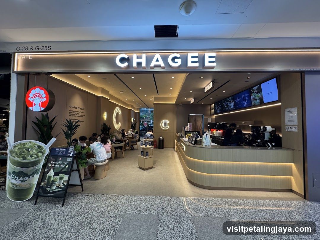 The exterior of a Chagee Tea Bar, a bubble tea shop. The shop has a modern design with a light wood counter and seating area. The large illuminated sign above the entrance reads "CHAGEE". There is a circular sign with a woman's face on the left side of the entrance. Inside, customers are seated at tables, and staff are visible behind the counter. A large cutout of a matcha drink is placed outside the entrance.