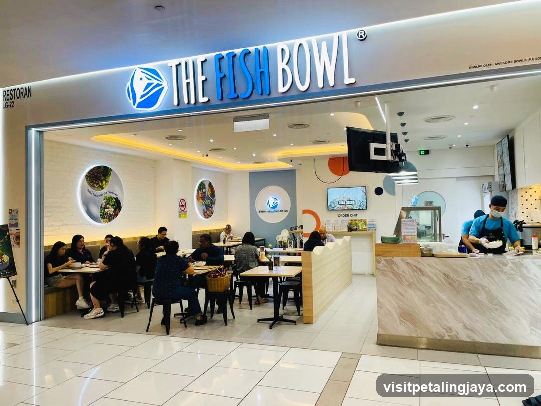 The exterior of a restaurant called The Fish Bowl. The restaurant has a bright, modern interior with customers seated at tables. A staff member in a blue apron and mask is preparing food behind a counter. The restaurant's logo and name are prominently displayed above the entrance.