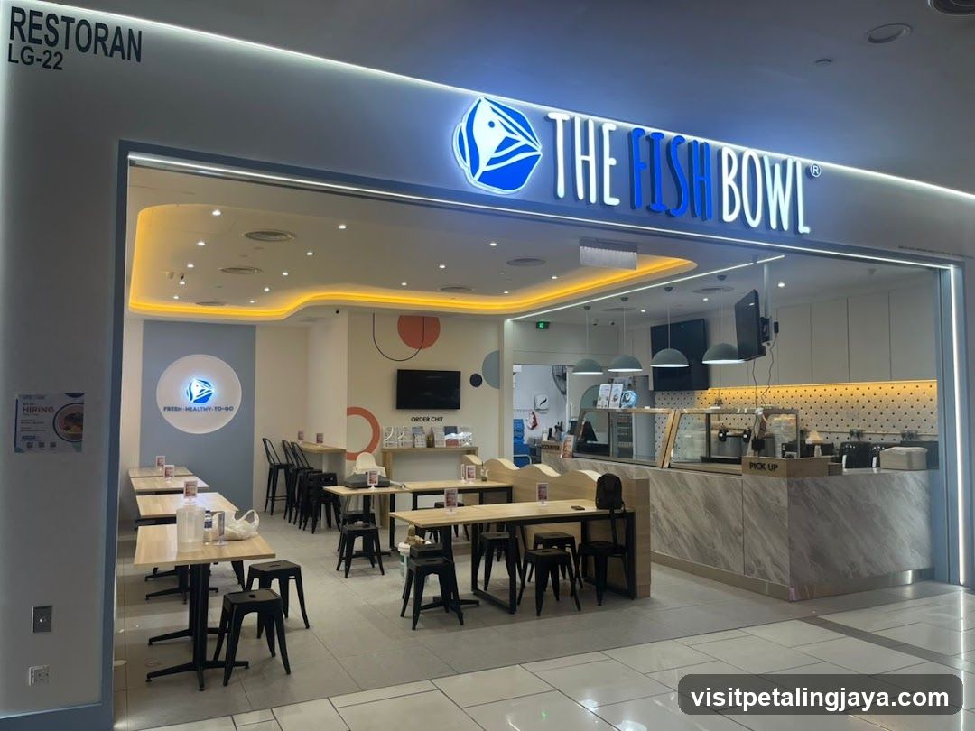 The exterior of a restaurant called The Fish Bowl, with its name prominently displayed in blue neon letters above the entrance. The interior is visible, showing tables and chairs, a counter area with a 'Pick Up' sign, and decorative elements. A 'Hiring' poster is visible on the left side of the entrance.