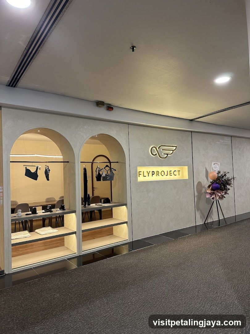 The exterior of a store with a modern, minimalist design. The facade features arched display windows showcasing athletic wear, such as bras and leggings. Above the windows, a prominent sign reads "FLYPROJECT" in illuminated golden letters, accompanied by a stylized wing logo. To the right of the sign, a floral arrangement with balloons adds a festive touch. The interior is visible through the windows, with shelves displaying folded items and racks holding garments.