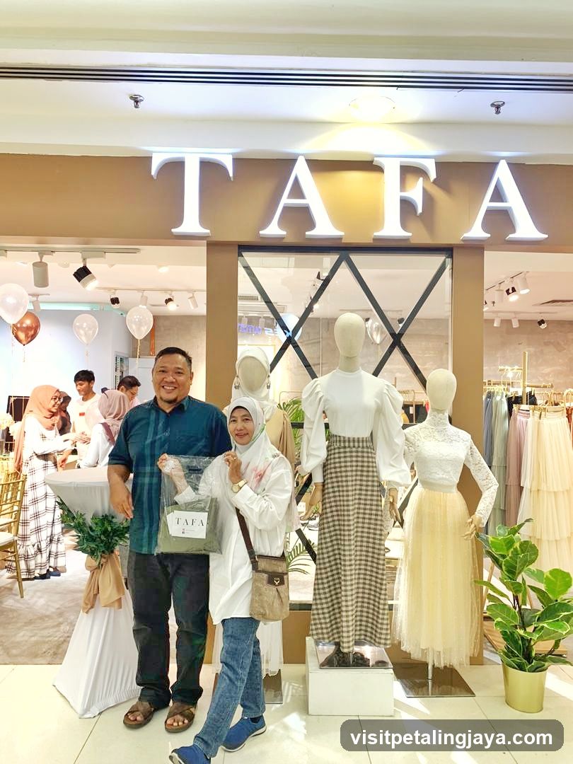 A couple stands in front of a store with the name "TAFA" displayed in large illuminated letters above the entrance. The store appears to be a clothing boutique, with mannequins dressed in fashionable attire visible through the glass storefront. Balloons add a festive touch to the scene.