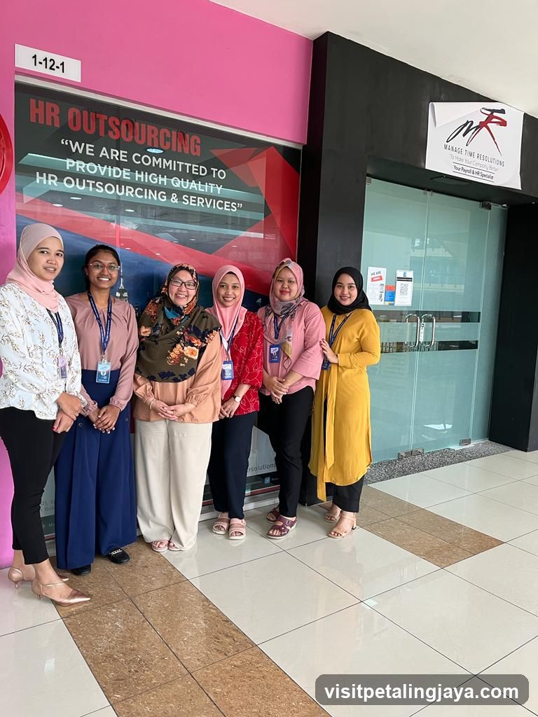 A group of six women stand in front of an office entrance. The wall to the left is pink and has a sign that reads "1-12-1" and "HR OUTSOURCING 'WE ARE COMMITTED TO PROVIDE HIGH QUALITY HR OUTSOURCING & SERVICES'". The entrance to the right is a glass door with a sign above it that says "MANAGE TIME RESOLUTIONS To Make Your Company Fee Your Payroll & HR Specialist".