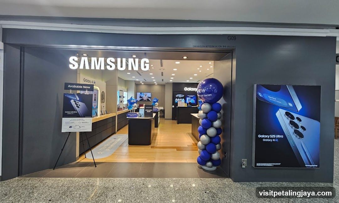 The exterior of a Samsung store with the Samsung logo prominently displayed above the entrance. Inside, various Samsung products are visible on display shelves and counters. Promotional materials, including posters and balloons, are placed outside and inside the store.