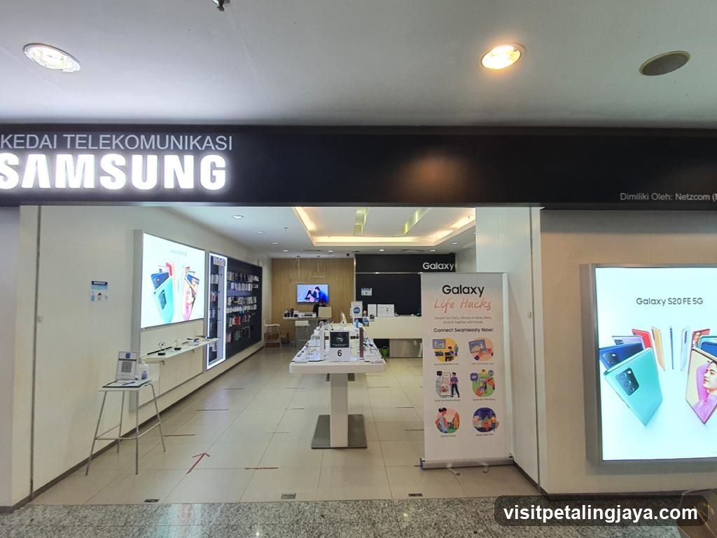 The exterior of a Samsung telecommunications store. The store has a large illuminated sign that reads "KEDAI TELEKOMUNIKASI SAMSUNG". Inside, display shelves are stocked with phones and accessories. A banner on the right advertises "Galaxy Life Hacks" and "Galaxy S20 FE 5G".