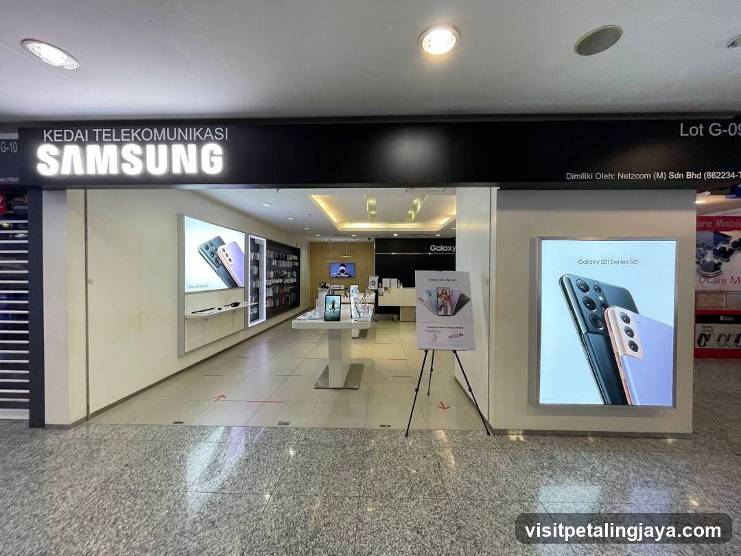The exterior of a Samsung telecommunication store. The store has a large illuminated sign that reads "KEDAI TELEKOMUNIKASI SAMSUNG". Inside, various Samsung phones and devices are displayed on shelves and stands. A large advertisement for the "Galaxy S21 Series 5G" is visible on the right side of the storefront.