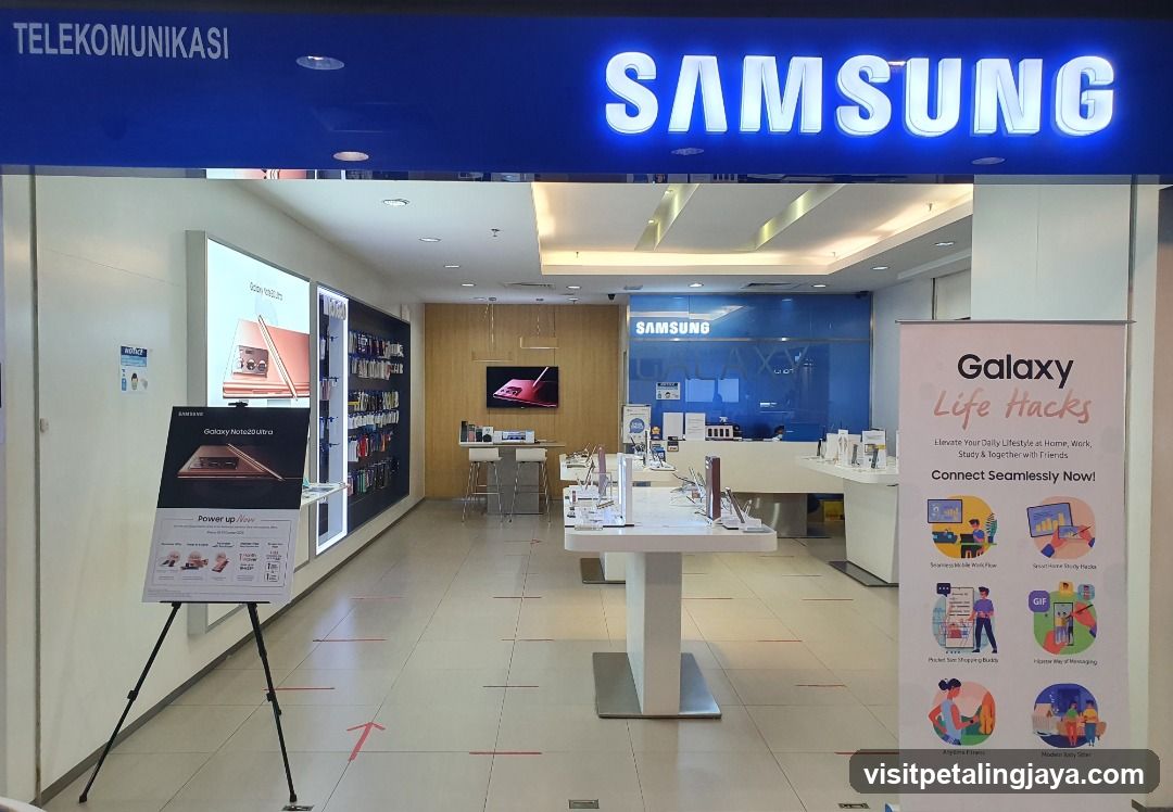 The exterior of a Samsung electronics store. A large illuminated "SAMSUNG" sign is visible above the entrance. Inside, display tables with phones and accessories are arranged. A promotional banner for "Galaxy Life Hacks" stands to the right.