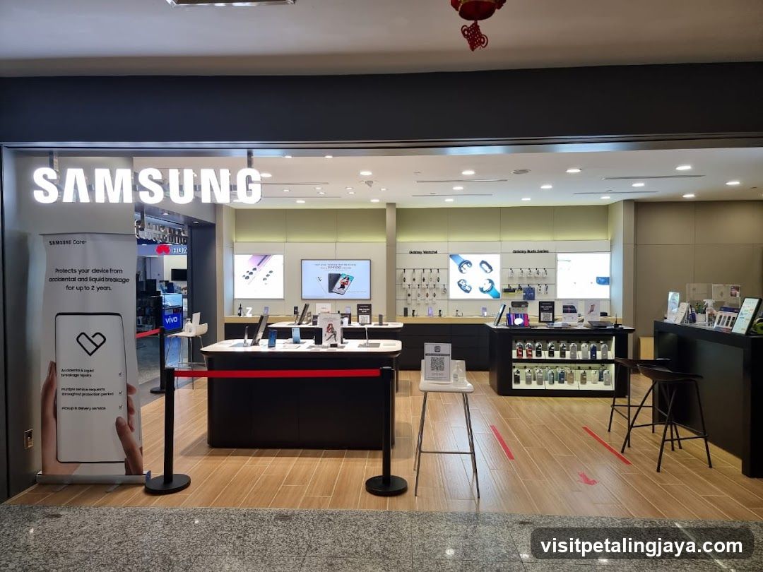 The exterior of a Samsung store with the large illuminated "SAMSUNG" logo above the entrance. Inside, various Samsung products like phones and watches are displayed on shelves and counters. A banner on the left advertises "Samsung Care" for device protection.