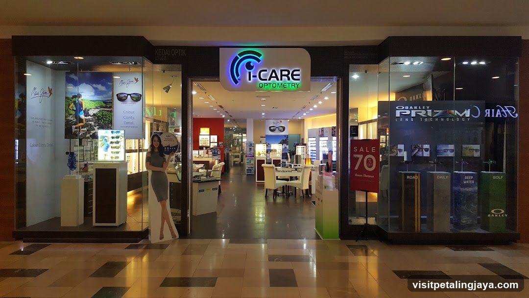 The image shows the exterior entrance of an optometry store named 'I-CARE OPTOMETRY'. The shop has a modern facade with large glass windows and doors. Inside, there are display counters, chairs, and various optical products visible. A sign on the window advertises a sale with 70% off. There is also branding for Ray-Ban and Oakley sunglasses displayed in the window.