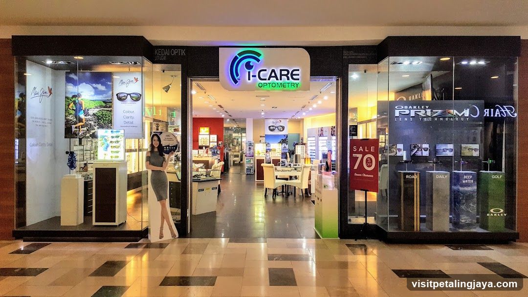 The image shows the exterior entrance of an optometry store named 'I-CARE OPTOMETRY'. The storefront features large glass windows and doors, with a prominent sign above the entrance displaying the store's logo and name. Inside, there are various displays of eyewear products, including sunglasses and glasses. A cutout figure stands near the entrance, and promotional posters advertise different brands like Oakley and Prizm technology. There is also a sale sign indicating discounts on certain items.