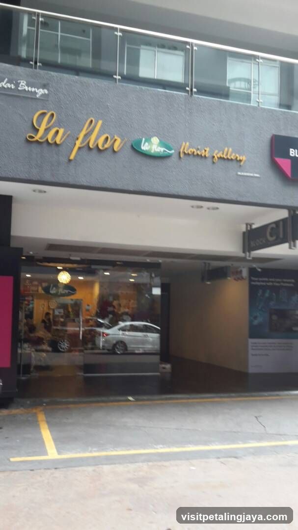 The image shows the storefront of La Flor Flower Gallery, featuring a modern facade with glass windows and metal railings above. The shop sign is prominently displayed in gold lettering on a dark background. Inside, there are visible displays of flowers and plants, along with customers browsing.