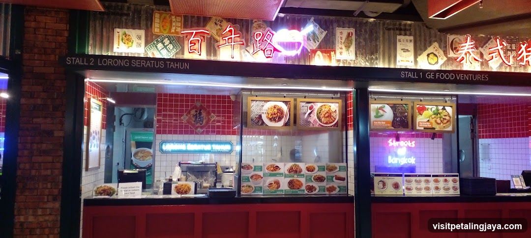 A food stall with red and white tiled walls, featuring illuminated signage in Chinese characters and English text indicating 'Stall 2 Lorong Seratus Tahun' and 'Stall 1 GE Food Ventures'. The display includes framed pictures of dishes, a neon sign reading 'Street Bangkok', and various menu boards showcasing food items.