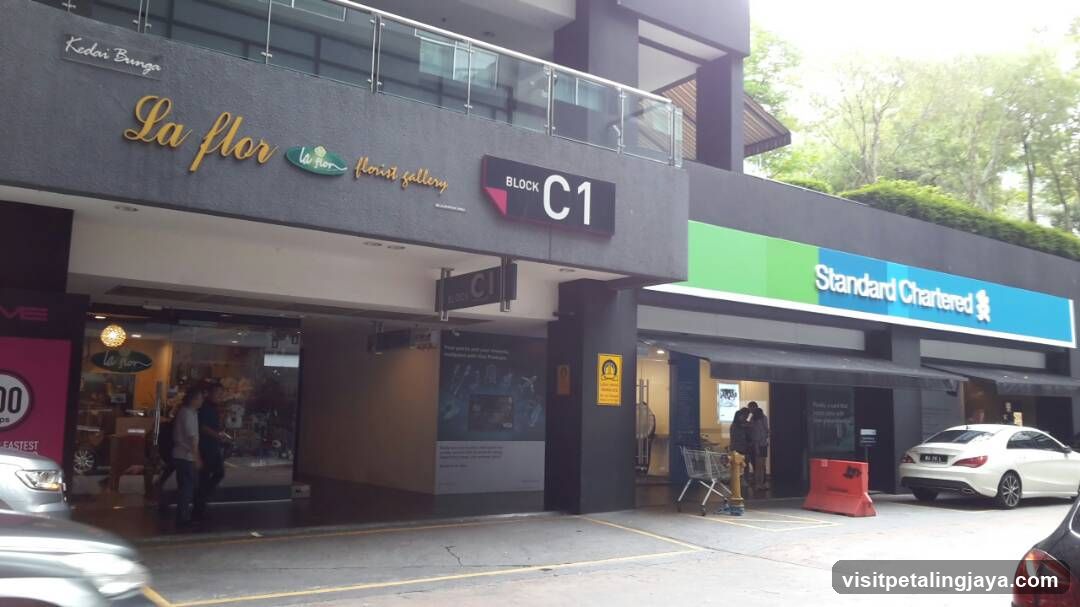 The image shows the exterior facade of a commercial building with two visible shop fronts. On the left, there is 'La flor flower gallery' with its name displayed in gold lettering on a dark grey wall above an entrance. A sign indicates this is Block C1. To the right, there is a branch of Standard Chartered bank with its logo prominently displayed on a blue and green sign. The ground level includes glass doors for La flor and a covered walkway leading to the bank entrance. There are parked cars nearby and some people standing near the entrances.