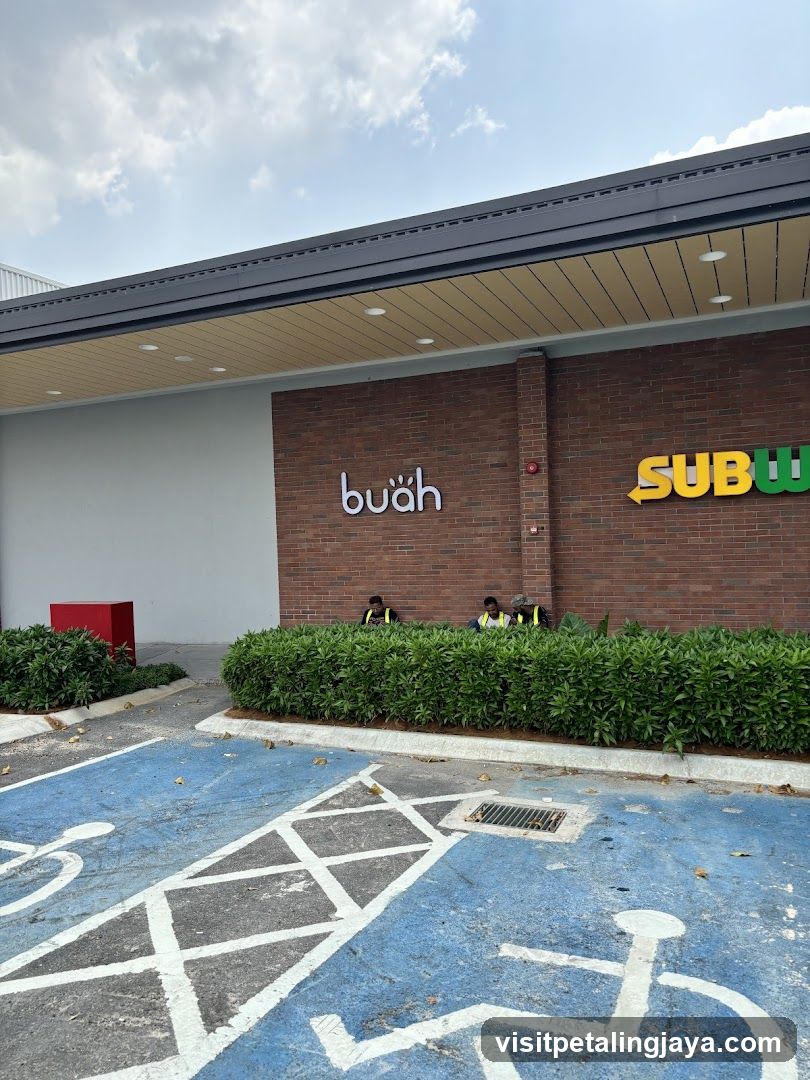The exterior of a building with two businesses, Subway and Buah. The Subway sign is yellow and green, and the Buah sign is white. There are three people sitting behind a hedge in front of the businesses. The foreground shows a blue parking space with white markings for a wheelchair and a bicycle.
