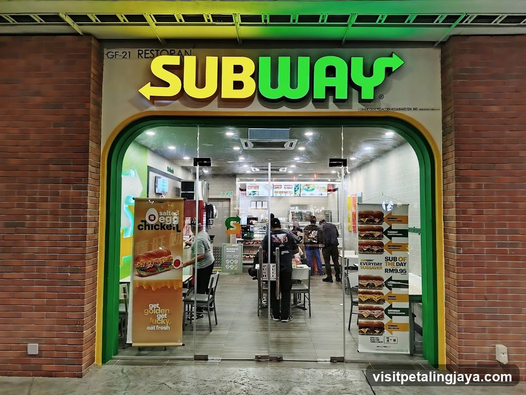 The exterior of a Subway restaurant with a large, illuminated Subway logo above the entrance. The entrance is made of glass doors, and through them, customers and staff can be seen inside. There are promotional banners displayed on either side of the entrance, one advertising a "Salted Egg Chicken" sandwich and another showcasing daily sandwich specials with a "Subsaver" price of RM9.95.