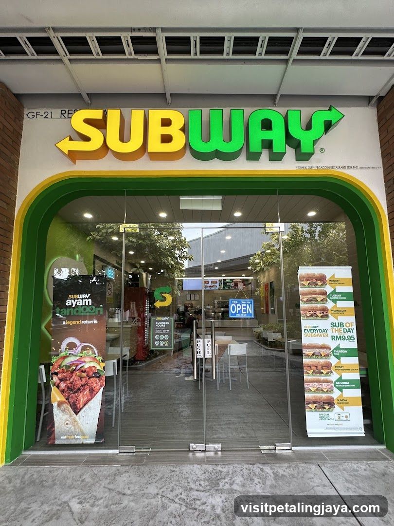 The exterior of a Subway restaurant with its prominent yellow and green signage above a glass entrance. A large promotional poster for "Subway Ayam Tandoori" is visible on the left glass door, and a "Sub of the Day" menu is displayed on the right. The interior of the store, including counters and seating, is visible through the glass doors. An "Open" sign is displayed on the door.