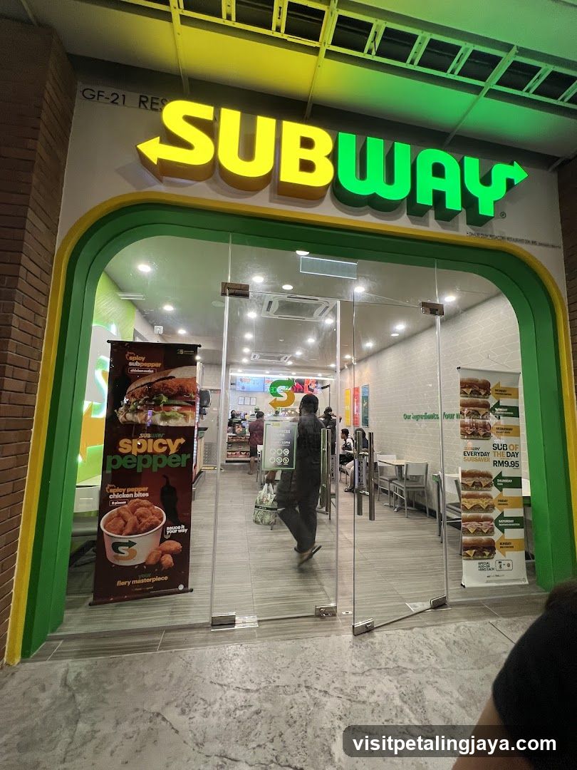 The exterior of a Subway restaurant with its prominent yellow and green signage above the entrance. The glass doors reveal the interior with tables and customers. A promotional banner for "Spicy Pepper Chicken Bites" is displayed on the left, and a "Sub of the Day" menu is on the right.