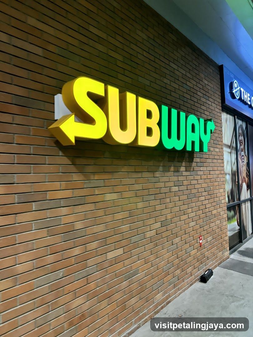 A large, illuminated Subway sign is mounted on a brick wall. The sign features the word "SUBWAY" in yellow and green, with a yellow arrow pointing to the left. To the right of the Subway sign, another storefront with a blue neon sign is partially visible.