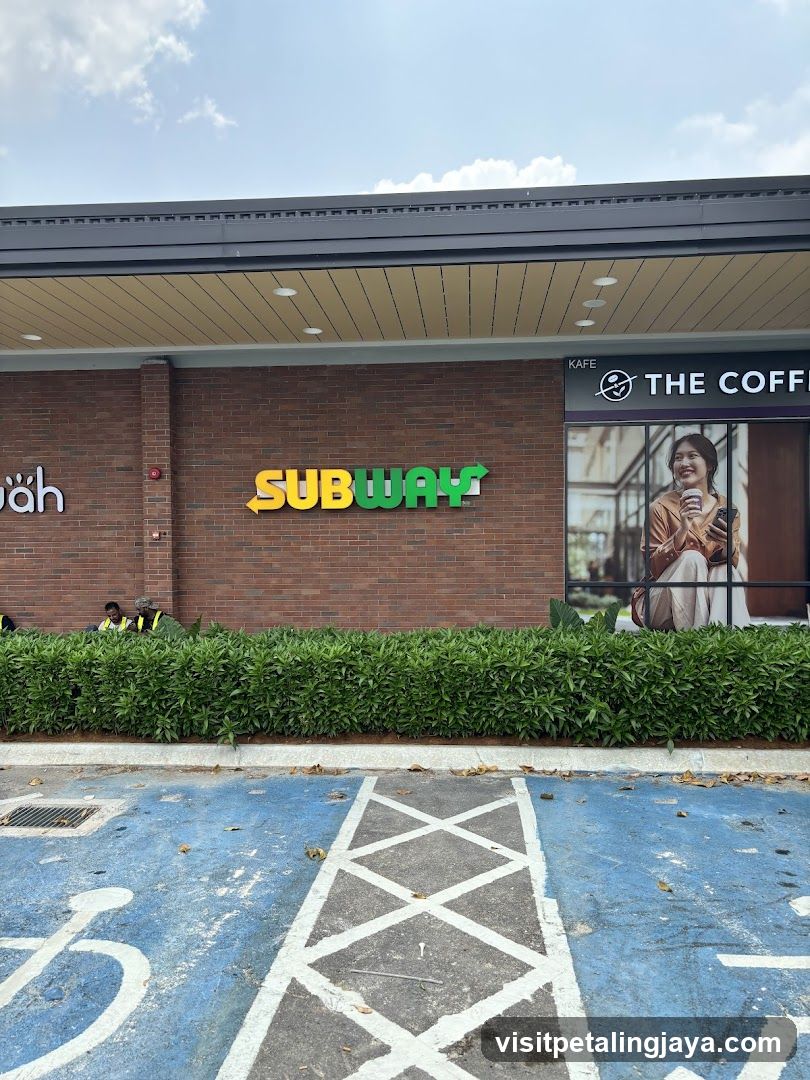 The exterior of a Subway restaurant with its iconic green and yellow logo prominently displayed on a brick wall. To the right, a portion of 'The Coffee Bean & Tea Leaf' storefront is visible, featuring a large advertisement with a woman holding a coffee cup. A neatly trimmed hedge runs along the base of the buildings, and a blue-painted parking area with white markings, including a wheelchair symbol, is in the foreground.