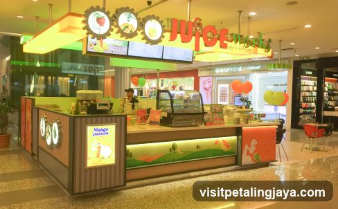 A brightly lit juice bar with a colorful facade and prominent signage. The counter has a display case with desserts and a blender. A staff member is visible behind the counter. The shop is located in a mall, with other stores visible in the background.