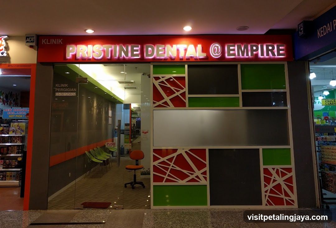 The exterior of a dental clinic with a prominent illuminated sign that reads "PRISTINE DENTAL @ EMPIRE". The entrance is glass, revealing a waiting area with green chairs and an orange swivel chair. The facade features a modern design with panels of green, red, and grey.