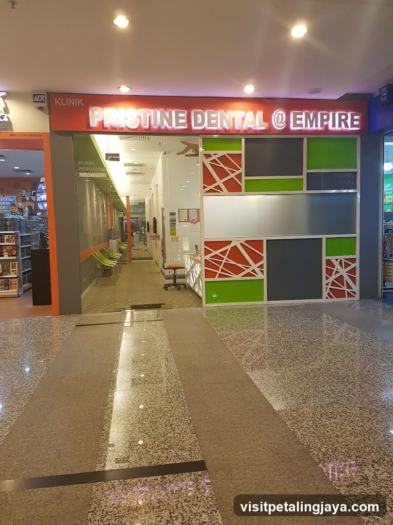 The exterior of a dental clinic with a prominent sign that reads "KLINIK PRISTINE DENTAL @ EMPIRE". The entrance is visible, with a hallway leading inside where chairs and a reception desk can be seen. The facade features a modern design with green, grey, and red geometric patterns.