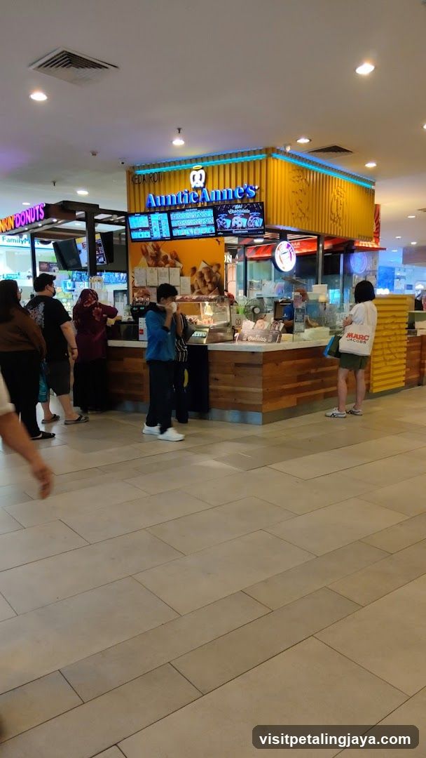 A view of the Auntie Anne's pretzel shop counter with customers ordering. The shop has a bright yellow facade with blue neon lighting. Digital menu boards display various pretzel options and prices.