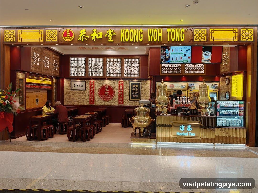 A brightly lit herbal tea shop with traditional Chinese decor. The shop front features a prominent sign with the name "KOONG WOH TONG" in yellow lettering. Inside, there are tables and stools for customers, a counter with large golden urns, and a display of bottled drinks. The walls are adorned with decorative panels and framed calligraphy.