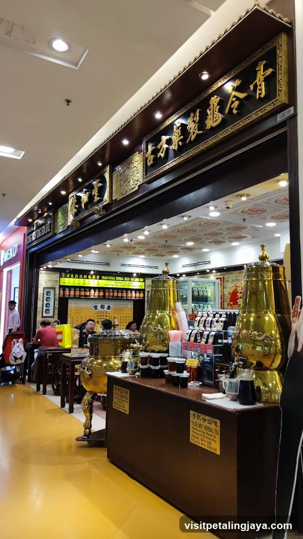 A view of the exterior of a shop with ornate golden signage and traditional Chinese architectural elements. Inside, customers are seated at tables, and a counter displays large golden urns, jars of herbal jelly, and cups. A sign advertises "TRADITIONAL HERBAL JELLY GULING GAO" and "PREVENTION IS BETTER THAN CURE." Another sign lists "A SE MEI HERBAL TEA" with benefits and prices.