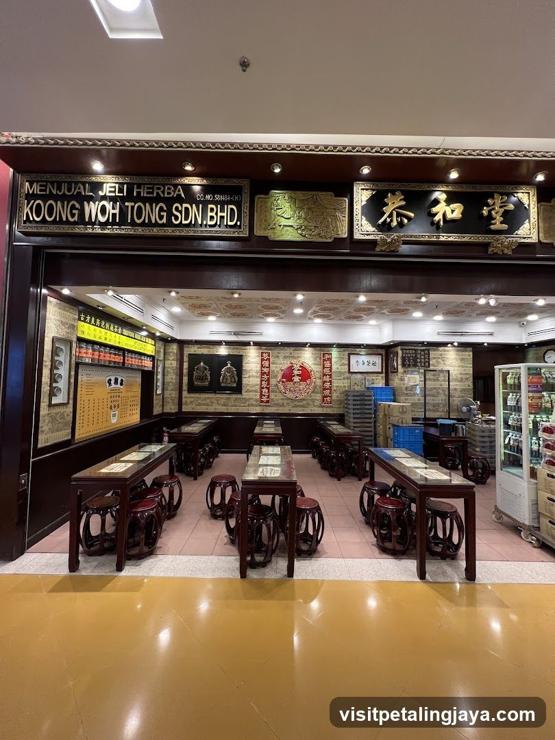 The exterior of a traditional herbal jelly shop with ornate signage and interior seating. The shop front features a prominent sign that reads "MENJUAL JELI HERBA KOONG WOH TONG SDN.BHD." and Chinese characters. Inside, there are rows of dark wood tables and stools, suggesting a seating area for customers. Shelves with products and decorative elements are visible along the walls.