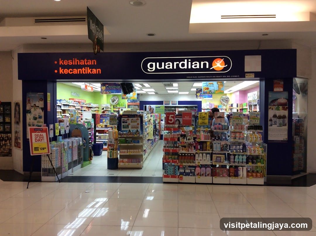 The image shows the entrance of a Guardian pharmacy store, with its name and logo prominently displayed above the doorway. The sign also includes the words 'kesihatan' (health) and 'kecantikan' (beauty). Inside, shelves are stocked with various health and beauty products, including personal care items, supplements, and household goods. A promotional standee is visible near the entrance.