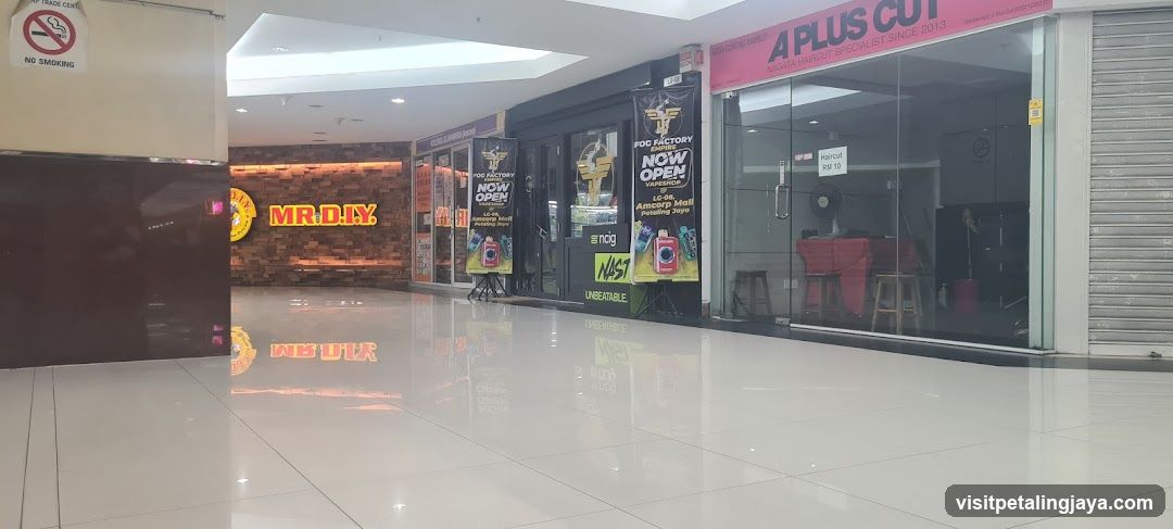 The image shows a brightly lit indoor shopping corridor with several storefronts visible. On the left, there is an entrance to 'MR.DIY' with its distinctive yellow and red signage on a brick-patterned wall. Adjacent to it are other shops including one advertising 'NOW OPEN' promotions and another labeled 'A PLUS CUT', which appears to be a hair salon or barbershop. The floor is highly reflective white tile, enhancing the brightness of the space. A no-smoking sign is affixed to the upper-left corner of the frame. No menus or price lists are visible in this scene.
