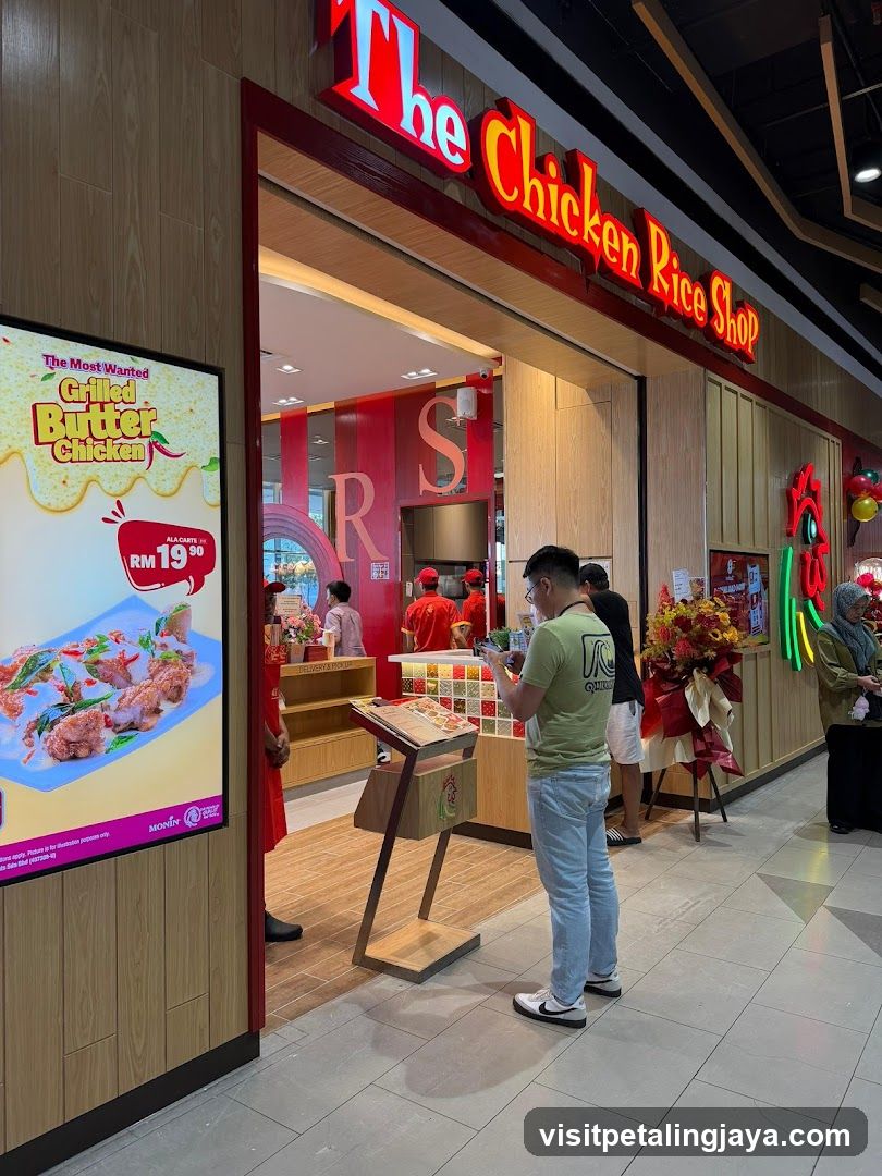 A storefront of The Chicken Rice Shop with visible signage and a digital menu board advertising Grilled Butter Chicken at RM19.90. Staff in red uniforms are inside, and customers are standing near the entrance.