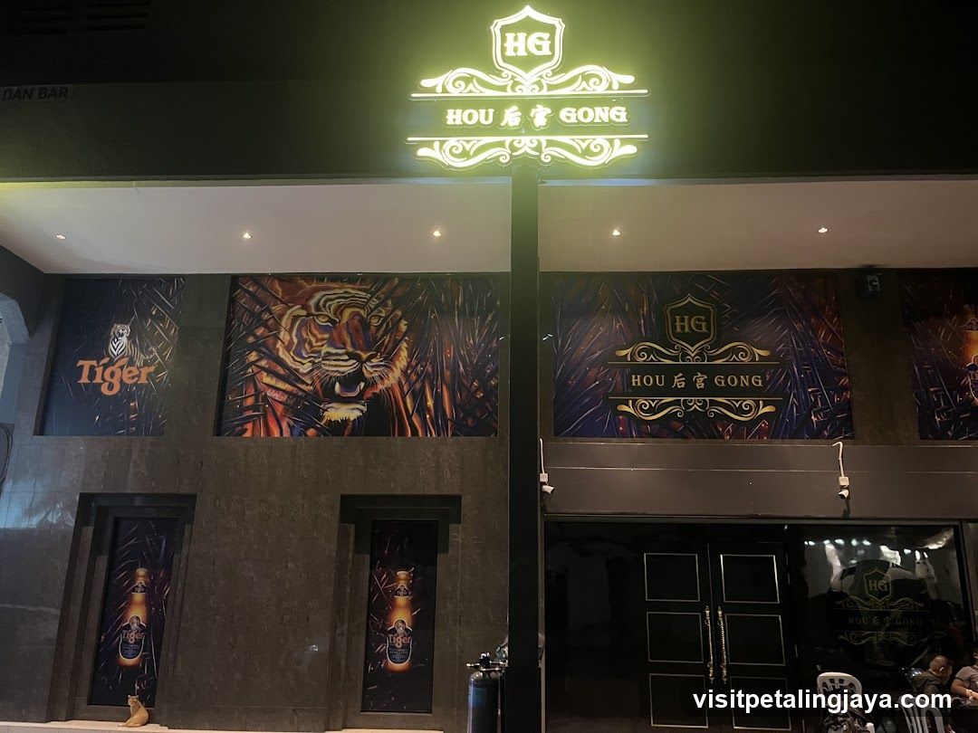 Night shot of a bar entrance with illuminated signage reading 'HG HOU 后宫 GONG'. The facade features tiger-themed artwork, including a large mural and branded posters. Two vertical banners display beer bottles near the doors. A person sits in the glass-enclosed seating area to the right.