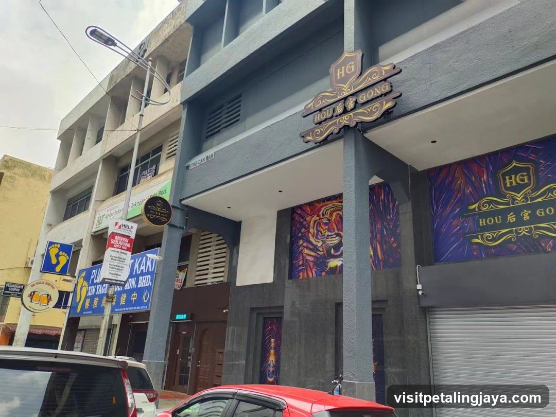 A multi-story building with a prominent 'HOU HOU GONG' sign above the entrance. The facade features decorative murals and several shop signs, including one for 'PLUMBER' and another partially visible sign. A red car is parked in front.