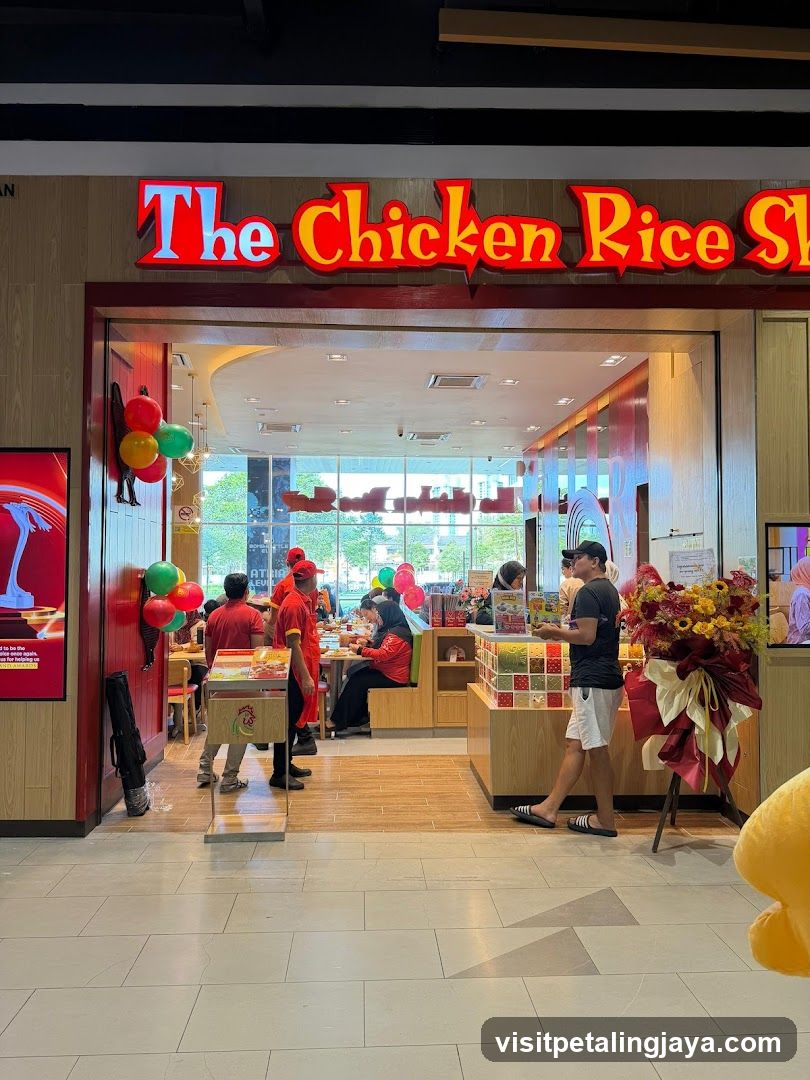 A brightly lit storefront with a red and yellow illuminated sign reading 'The Chicken Rice Sh'. The entrance is wide, revealing an interior with customers seated at tables and staff in red uniforms. Decorative balloons (red, green, yellow) are clustered near the left door frame. A large floral arrangement stands to the right of the entrance. Inside, wooden flooring extends into the dining area with glass windows showing daylight outside. A person in casual clothing walks past the counter on the right side.