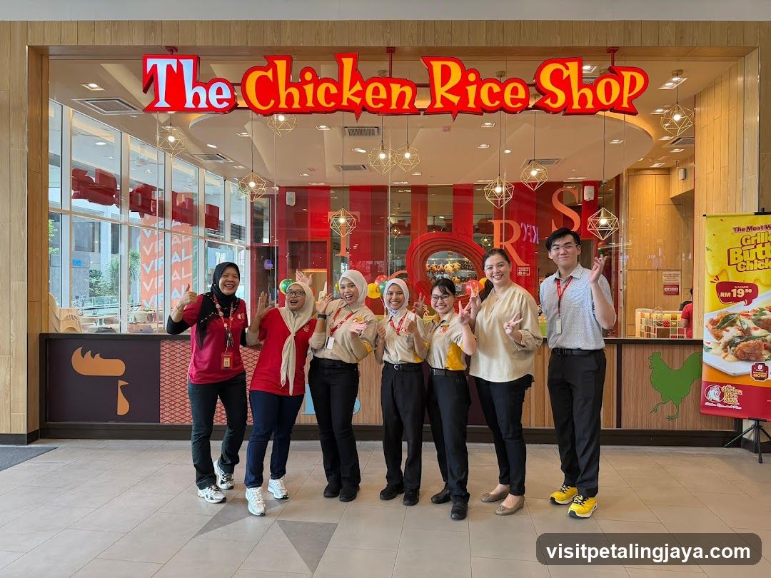 A group of seven staff members pose in front of The Chicken Rice Shop storefront, which features a prominent red and yellow sign above the entrance. Inside, hanging pendant lights illuminate the dining area with red accents.