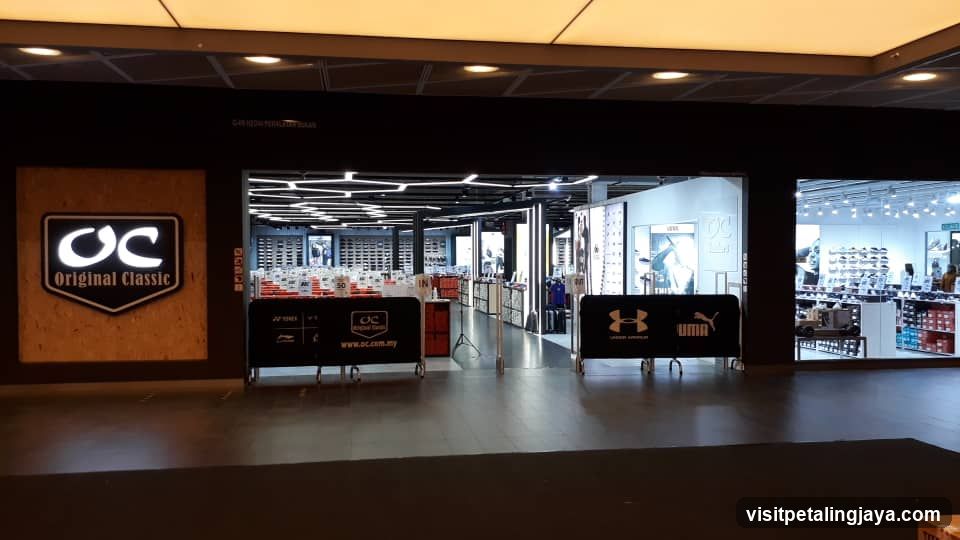 The image shows the storefront of a shop named 'Original Classic', featuring a prominent illuminated logo on a wooden panel to the left. The entrance is wide, leading into a brightly lit interior with multiple shoe displays and racks. Two black barriers with brand logos (Under Armour and Puma) are placed at the entrance, partially blocking access. The ceiling has recessed lighting and geometric patterns.