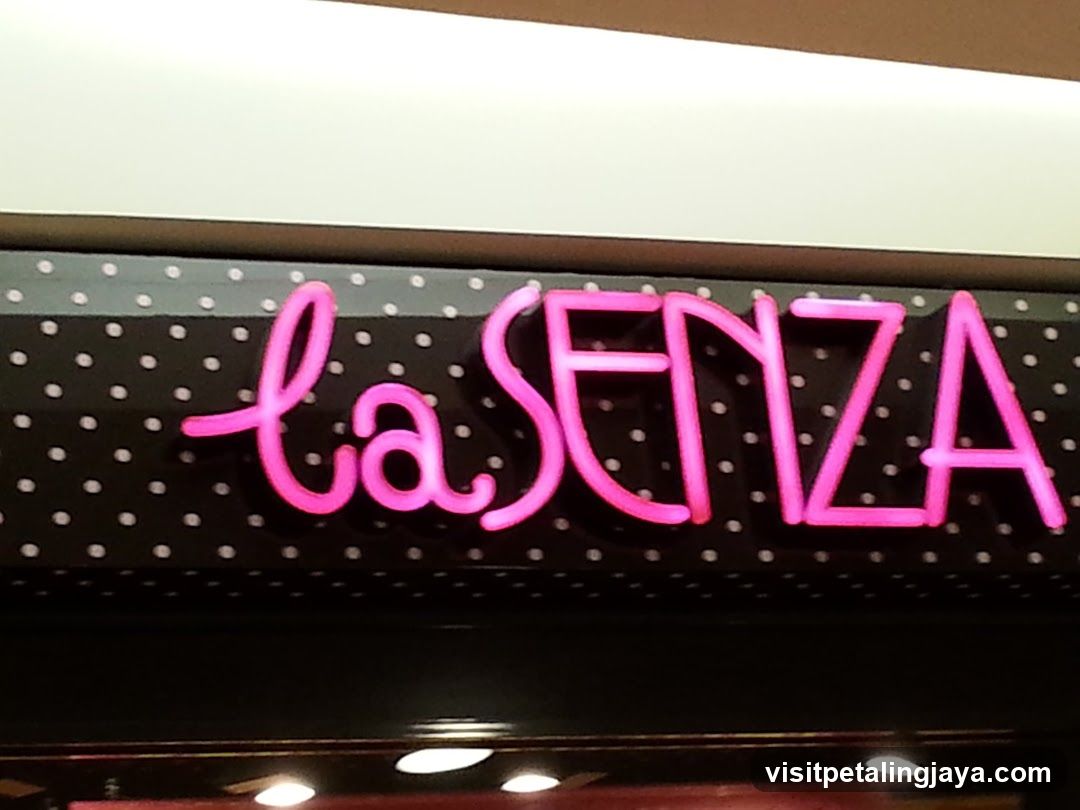A close-up view of a brightly lit neon sign displaying the brand name 'la SENZA' in pink cursive and block letters against a black background with white polka dots. The sign is mounted on what appears to be an exterior storefront facade, likely above or near the entrance of a retail store.