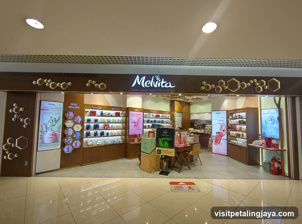 The image shows the storefront of a Melvita shop, featuring a prominent sign with the brand name 'Melvita' and its logo. The entrance is flanked by illuminated hexagonal designs on the walls. Inside, there are shelves stocked with various products, including skincare items and other goods. A promotional display table is visible near the entrance, along with posters advertising specific products. The floor is tiled, and the overall ambiance suggests a clean and organized retail environment.