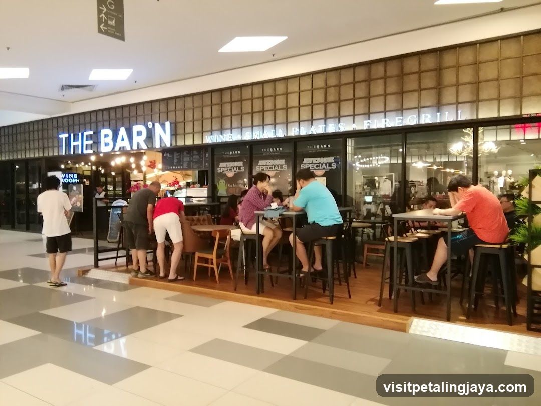 A photo of the physical storefront facade of 'THE BAR'N', a restaurant located in what appears to be a shopping mall. The building features large glass windows and doors, with illuminated signage reading 'THE BAR'N' above the entrance. Below this, additional signs indicate offerings like 'WINE', 'BAR', 'GRILL', 'PASTAS', and 'FIREGRILL'. Several customers are seated at high tables outside on wooden flooring, while others stand near the entrance. A person wearing a white shirt is visible near the left side of the frame.