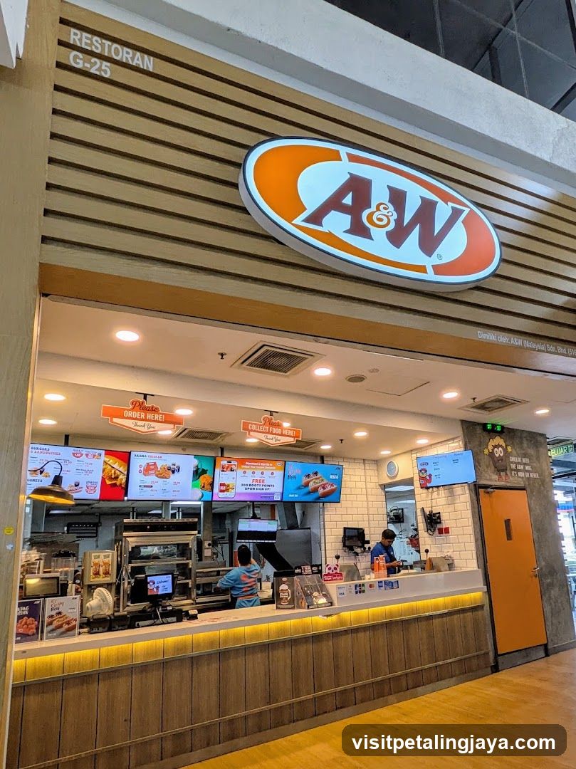 The image shows the storefront of an A&W restaurant, with its logo prominently displayed above the entrance. The sign reads 'RESTORAN G-25' and features a wooden facade with horizontal slats. Inside, there are digital menu boards displaying various food items, along with staff members working behind the counter.