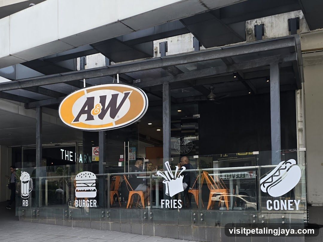 Photo of the physical storefront facade featuring a large A&W logo sign mounted above glass windows. Inside, customers are seated at orange chairs near the counter. Decorative decals on the window include icons labeled 'RB', 'BURGER', 'FRIES', and 'CONEY'. The building has an industrial-style metal frame structure with visible beams overhead.