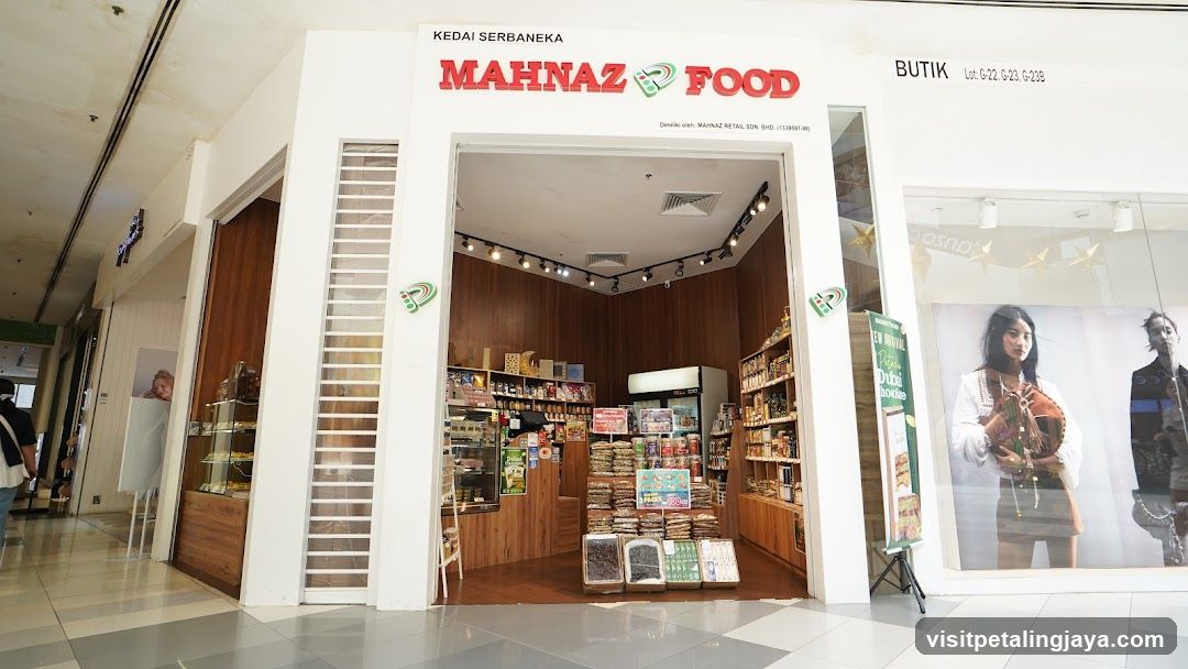 A brightly lit storefront with white facade and wooden interior accents. The sign reads 'KEDAI SERBANEA MAHNAZ FOOD' in red letters above the entrance. Inside, shelves display packaged goods, snacks, and beverages. A glass display case is visible on the left side of the store.