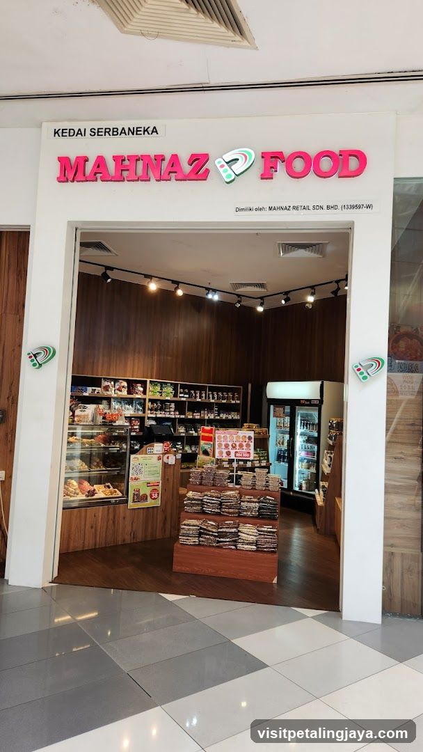 A photo of the physical exterior entrance of a shop named 'MAHNAZ FOOD' with visible signage above the doorway. The store interior shows shelves stocked with various products, including packaged goods and refrigerated items. A display stand in front holds neatly stacked packaged snacks or breads. Lighting fixtures are mounted on the ceiling inside.