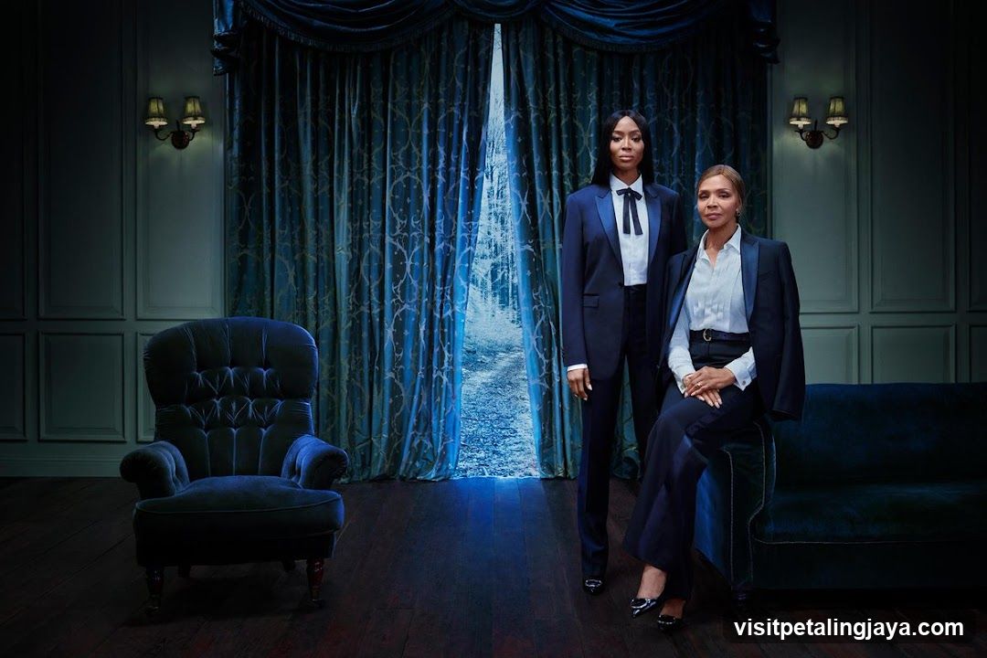 Two women in formal attire standing and sitting in a dimly lit room with blue curtains.