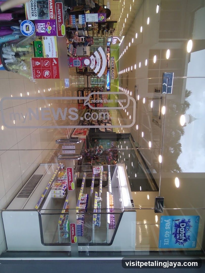 Photo of the physical exterior entrance of a convenience store or newsstand, viewed through glass doors. Visible signage includes 'myNEWS.com' on the door and various promotional stickers inside. The interior shows shelves stocked with products, including beverages and snacks, under bright lighting.