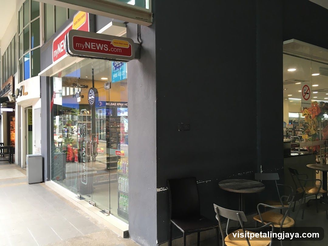 A shop front with a visible 'myNEWS.com' sign above the entrance. The store has glass doors and windows, displaying various products inside. There is an outdoor seating area with tables and chairs adjacent to the building.