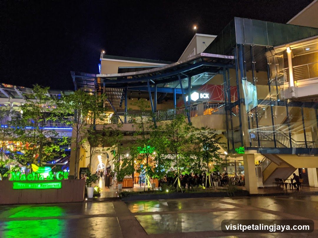 Night scene of a commercial complex with illuminated storefronts and greenery.