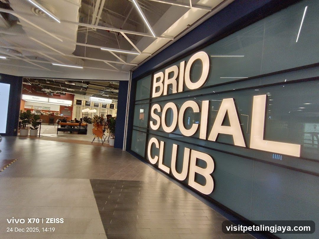 Photo of the storefront facade showing large illuminated signage reading 'BRIO SOCIAL CLUB' on a glass wall. Interior view visible through open entrance reveals modern seating, plants, and kitchen area in background.